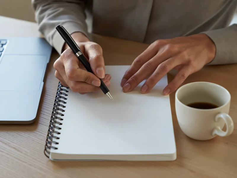 Illustration: Hände schreiben in ein leeres Notizbuch neben Laptop und Kaffeetasse